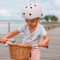 Fillette portant le casque vélo enfant Bebikeen XS rose pour les enfants de 2 à 6 ans,, assise sur son vélo avec panier en osier, en bord de mer.`