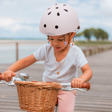 Fillette portant le casque vélo enfant Bebikeen XS rose pour les enfants de 2 à 6 ans,, assise sur son vélo avec panier en osier, en bord de mer.`