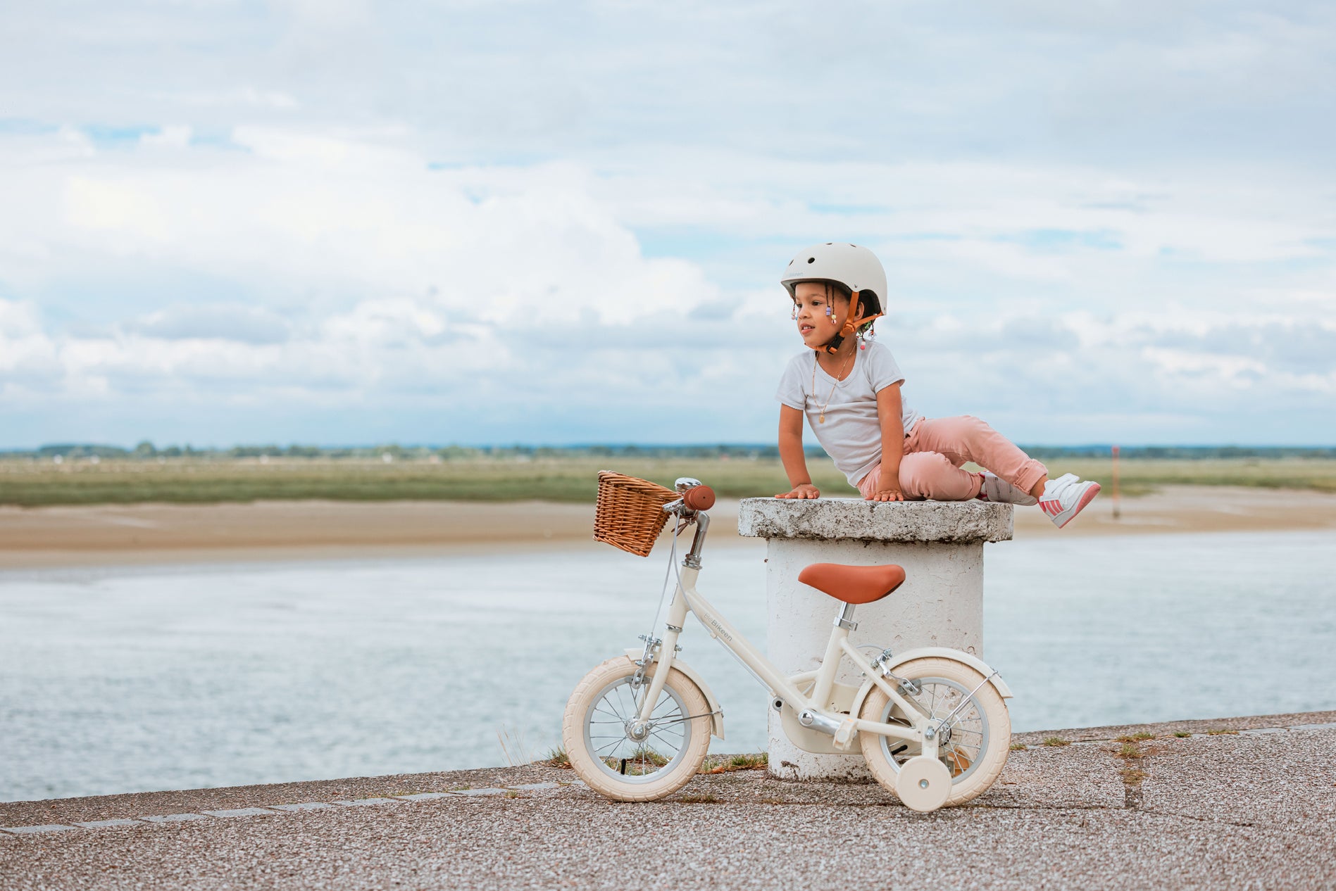 Petite fille assise sur un muret en bord de mer à côté de son vélo enfant 12 pouces BeBikeen crème avec panier en osier et stabilisateurs.