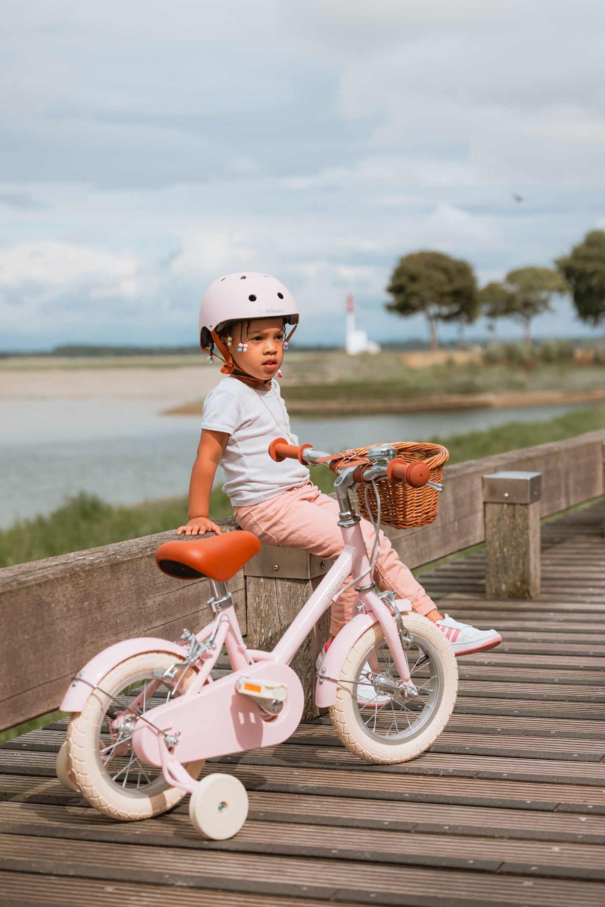 Fillette de 3 ans portant un casque rose Bebikeen à côté de son vélo 12 pouces rose avec panier en osier et roues stabilisatrices, au bord de la mer.