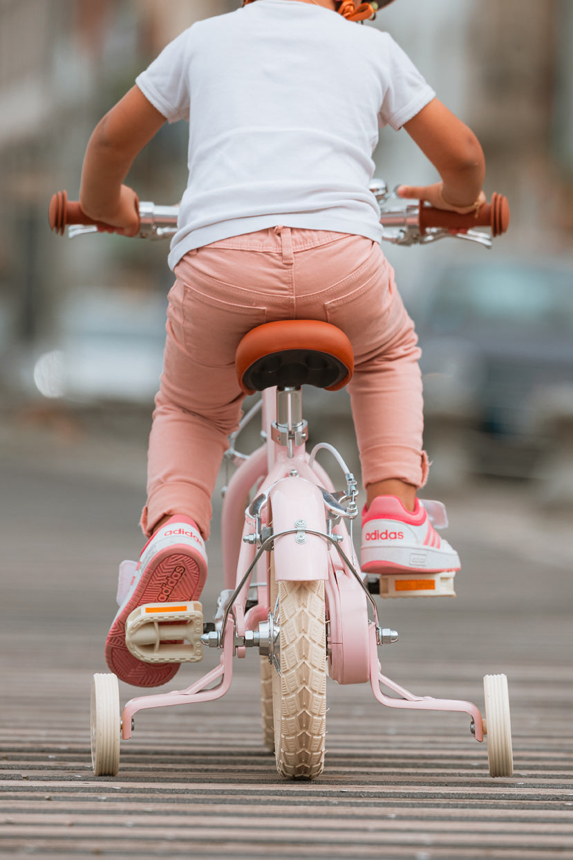 Enfant vue de dos pédale sur un vélo léger Bebikeen 12 pouces rose, équipé de roues stabilisatrices et d’une selle marron.