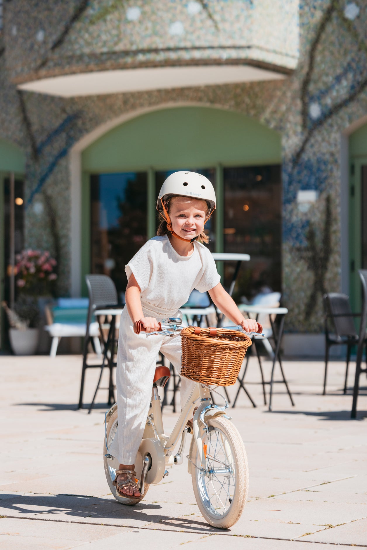Fillette portant un casque crème Bebikeen, pédale de face sur son vélo léger 16 pouces crème avec panier en osier, photo prise au Touquet.