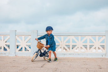 Petit garçon portant un casque bleu marine Bebikeen tient son vélo 12 pouces bleu marine avec panier en osier, au bord de la mer.