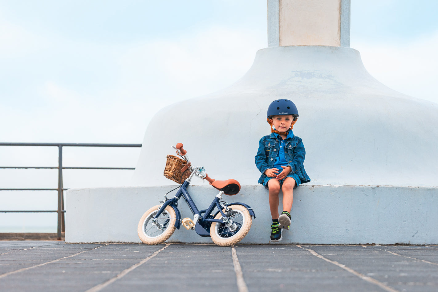 Garçon de 3 ans assis avec son casque bleu marine Bebikeen, à côté de son vélo léger enfant 12 pouces bleu marine avec panier en osier.