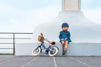 Garçon de 3 ans assis avec son casque bleu marine Bebikeen, à côté de son vélo léger enfant 12 pouces bleu marine avec panier en osier.