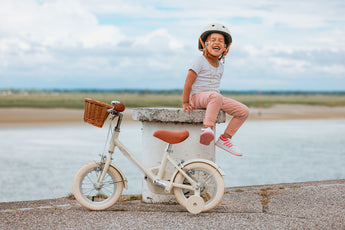 Fillette de 3 ans éclatant de rire, portant un casque de  vélo crème, assise près de son vélo enfant Bebikeen 12 pouces crème avec roues stabilisatrices et panier en osier.