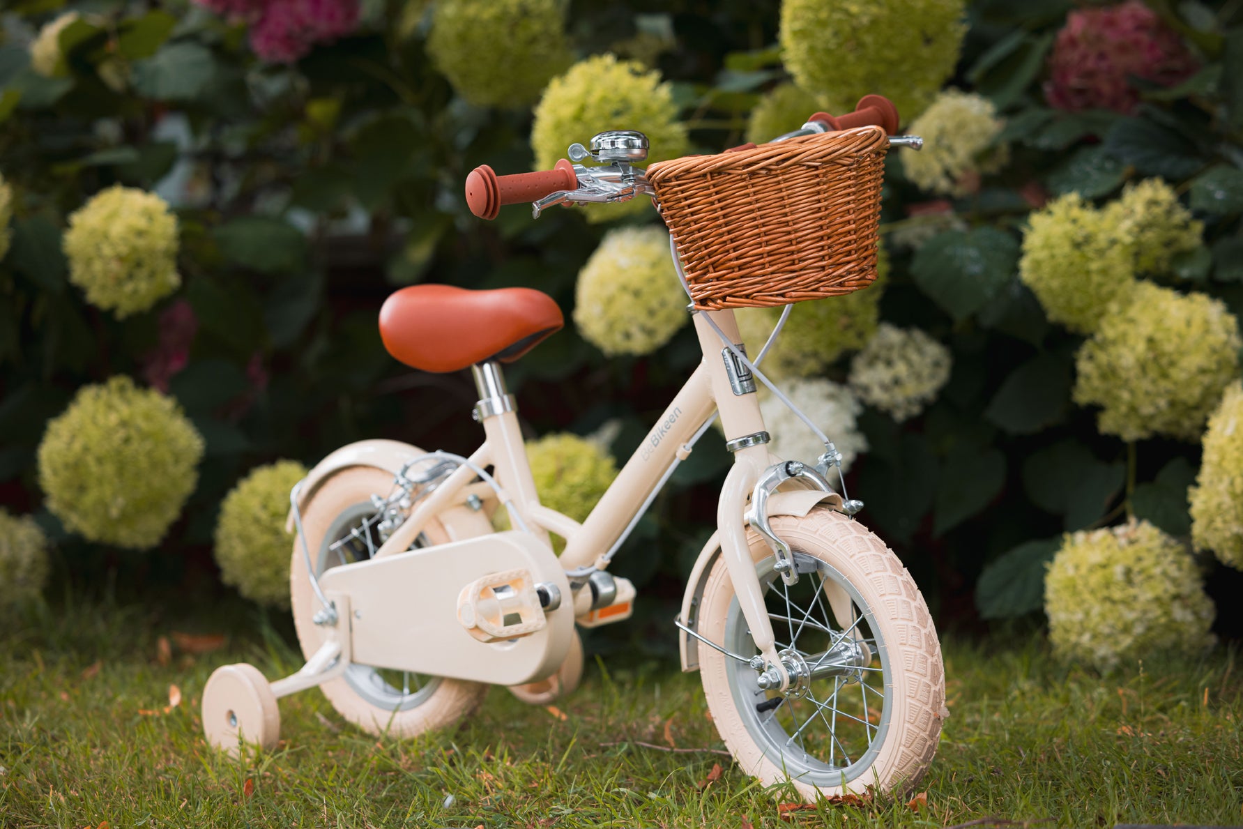 Vélo enfant Bebikeen 12 pouces crème, 2 à 4 ans, profil droit avec roues stabilisatrices et panier en osier, dans un jardin.