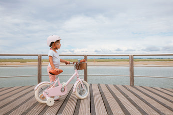 Fillette portant un casque rose Bebikeen, debout à côté de son vélo enfant 12 pouces rose avec panier en osier et stabilisateurs, regardant la mer au Touquet.
