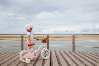 Fillette de 3 ans portant un casque rose, tenant son vélo léger Bebikeen 12 pouces rose avec stabilisateurs et panier en osier, en promenade au bord de l’eau.