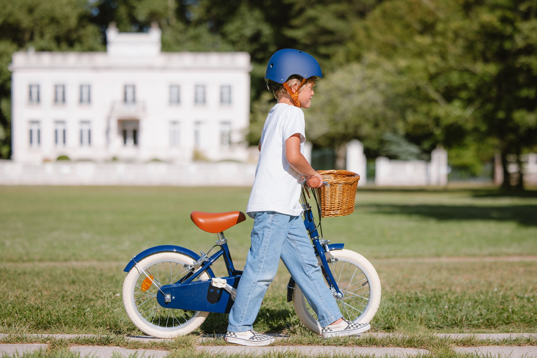 Garçon marchant à côté de son vélo 16 pouces Bebikeen bleu marine léger avec panier en osier et selle marron dans un parc.