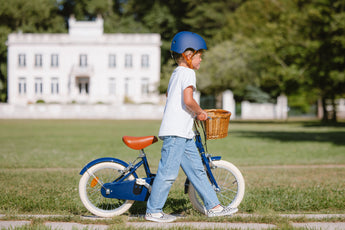 Garçon marchant à côté de son vélo 16 pouces Bebikeen bleu marine léger avec panier en osier et selle marron dans un parc.