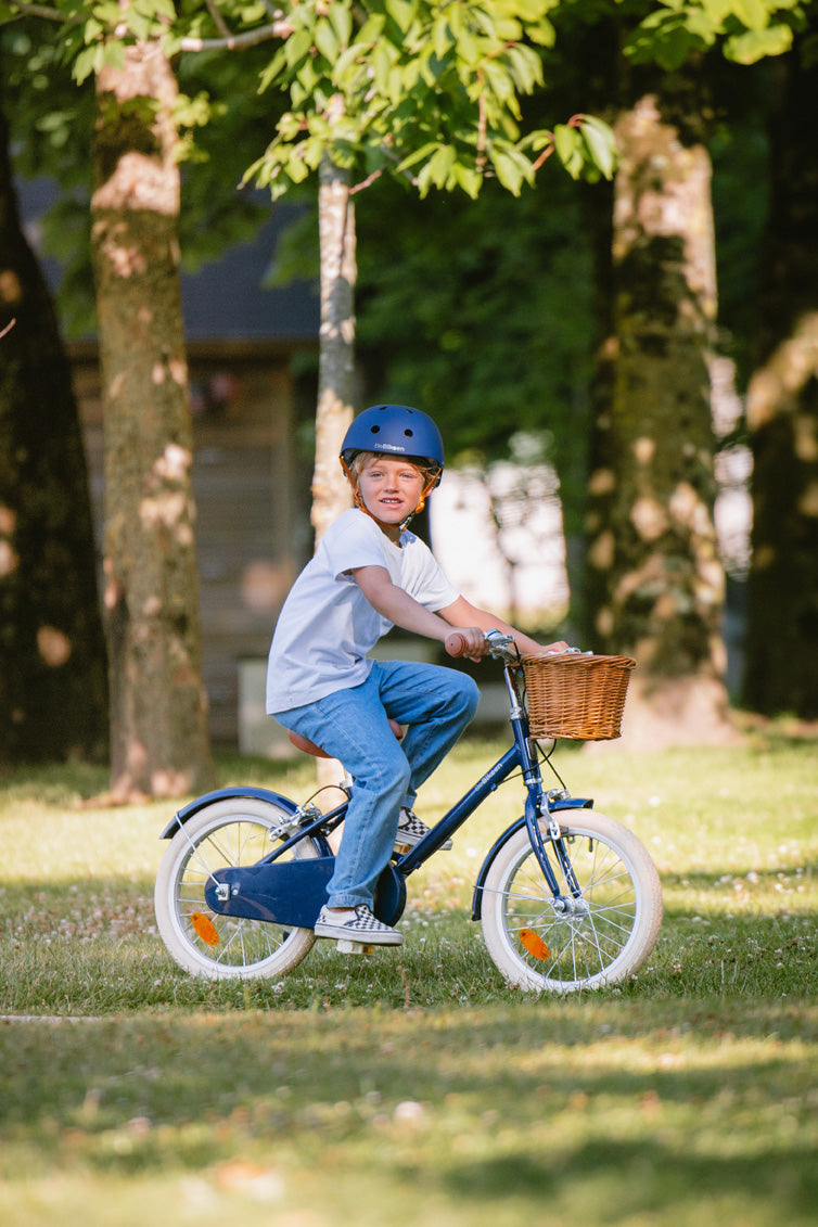 Garçon pédalant sur son vélo 16 pouces bleu marine BeBikeen avec panier en osier et une selle marron dans un de parc.