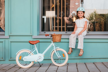 Fille de 6 ans assise à côté de son vélo léger Bebikeen 16 pouces bleu turquoise avec panier en osier et béquille, portant un casque bleu turquoise assorti.
