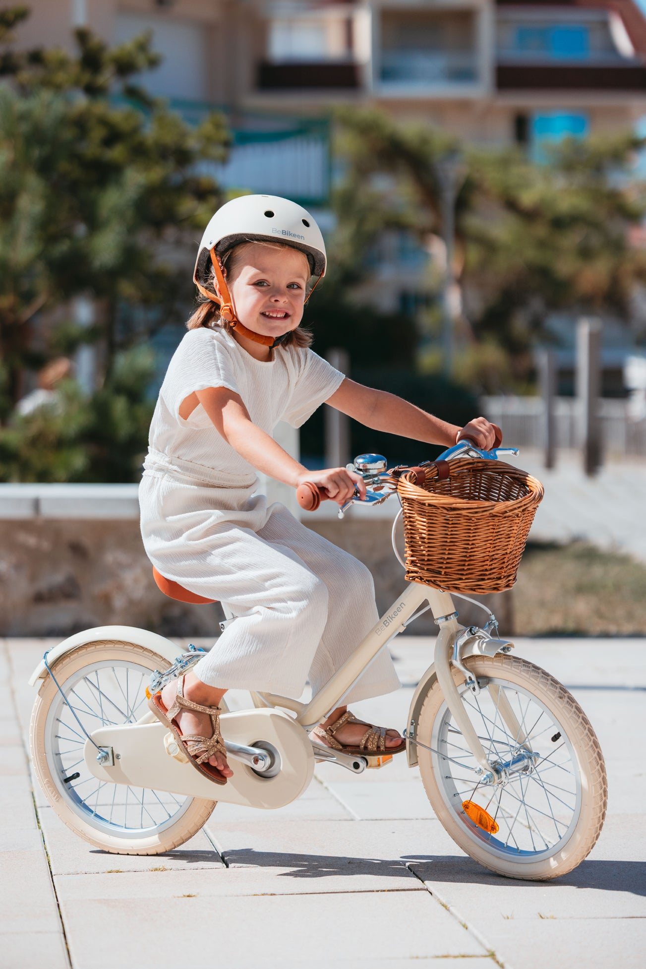 Fillette de 5 ans souriante portant un casque crème Bebikeen, pédale sur son élégant vélo enfant 16 pouces crème avec panier en osier et selle marron.