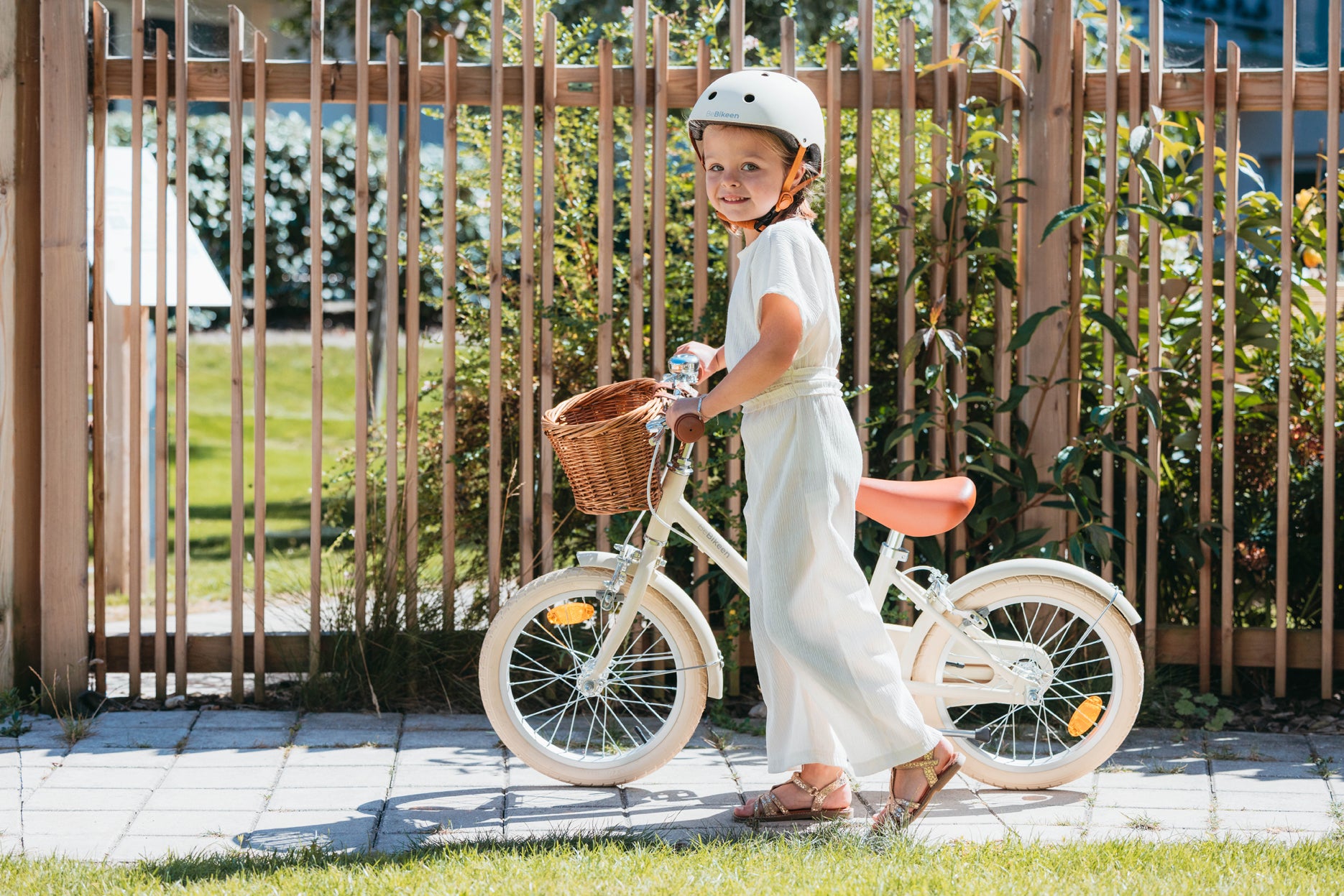 Fille 5 ans souriante, portant un casque crème Bebikeen, marche à côté de son vélo enfant 16 pouces crème avec panier en osier et selle marron au Touquet.