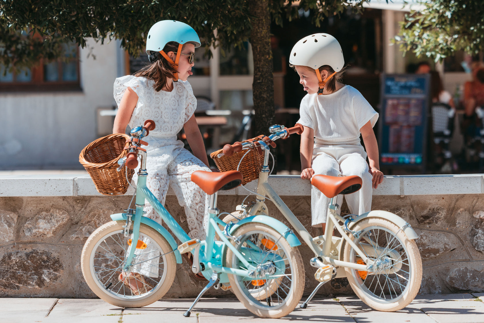 Deux fillettes s'amusent assises à côté de leur vélo Bebikeen 16 pouces, bleu turquoise et crème, équipés d’un panier en osier. Elles portent les casques assortis aux vélos.