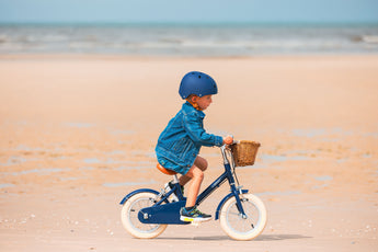 Garçon de 3 ans portant un casque bleu marine Bebikeen, pédale sur son vélo léger 12 pouces bleu marine avec panier en osier, sur la plage du Touquet