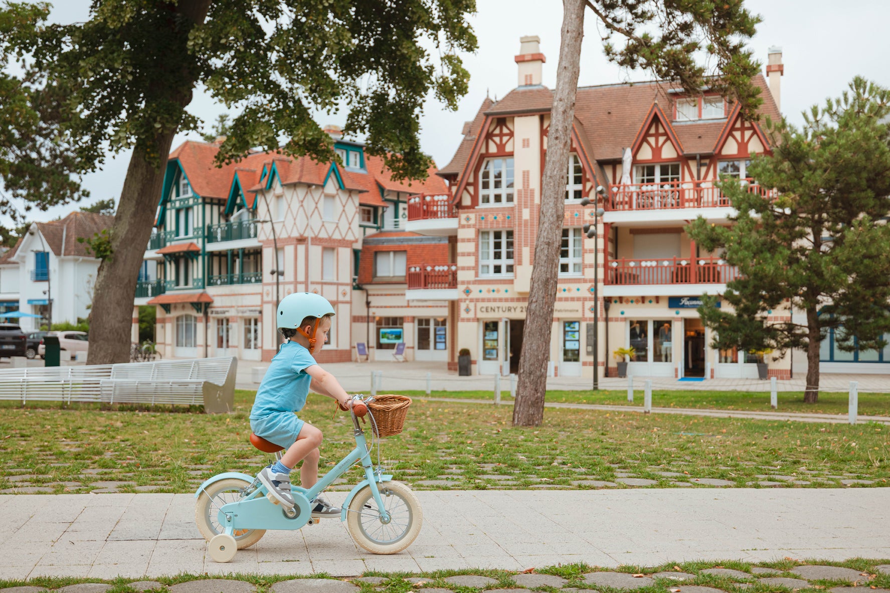 Garçon de 3 ans portant un casque bleu turquoise, pédalant sur un vélo Bebikeen 12 pouces bleu turquoise avec stabilisateurs et panier en osier. photo de  profil prise au Touquet.