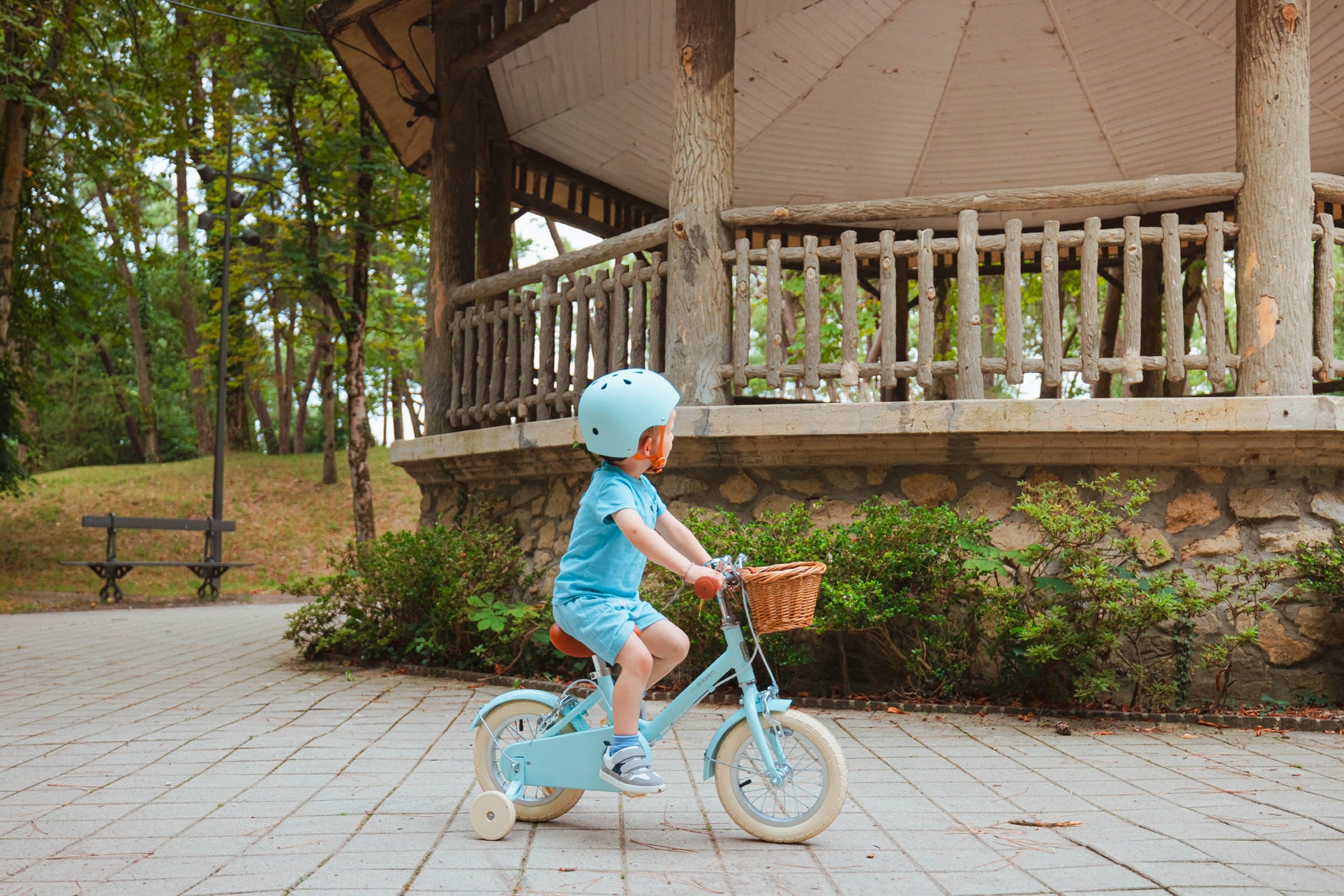 Garçon de 3 ans portant un casque bleu turquoise, pédalant sur un vélo enfant Bebikeen 12 pouces bleu turquoise avec stabilisateurs et panier en osier, dans un parc.