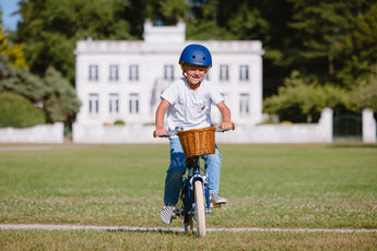 Garçon portant un casque bleu marine Bebikeen pédale sur son vélo léger 16 pouces bleu marine avec panier en osier, dans un parc au  Touquet.
