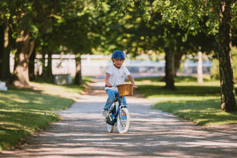 Garçon portant un casque bleu marine Bebikeen pédale sur son vélo 16 pouces bleu marine avec panier en osier, dans une allée de parc.