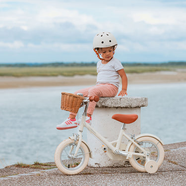 Fille de 3 ans assise à côté de son vélo enfant Bebikeen 12 pouces crème léger, 2 à 4 ans, profil avec panier et stabilisateurs, à la mer au Touquet.