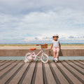 Fille de 3 ans à côté de son vélo léger Bebikeen 12 pouces rose, pour enfants de 2 à 4 ans, de profil avec panier, photo prise en bord de mer.
