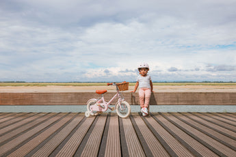 Fille de 3 ans à côté de son vélo léger Bebikeen 12 pouces rose, pour enfants de 2 à 4 ans, de profil avec panier, photo prise en bord de mer.