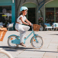 Jeune fille pédalant sur vélo enfant léger Bebikeen 16 pouces bleu turquoise avec casque assorti et panier en osier naturel, vue de profil.