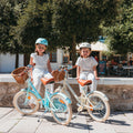 Deux filles assises à côté de leurs vélos Bebikeen 16 pouces bleu turquoise et crème, équipés de béquilles et de paniers en osier.