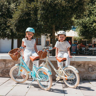 Deux filles assises à côté de leurs vélos Bebikeen 16 pouces bleu turquoise et crème, équipés de béquilles et de paniers en osier.