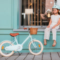 Fille assise à côté de son vélo Bebikeen 16 pouces bleu turquoise, pour enfant de 4 à 6 ans, équipé d’une béquille et d’un panier en osier.