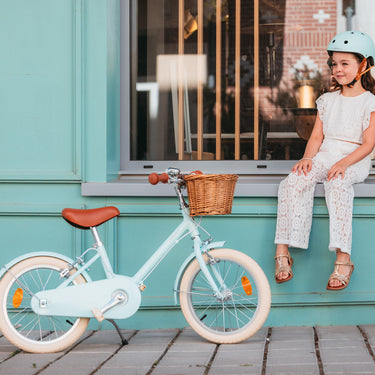 Fille assise à côté de son vélo Bebikeen 16 pouces bleu turquoise, pour enfant de 4 à 6 ans, équipé d’une béquille et d’un panier en osier.