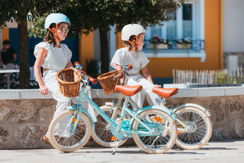 Deux fille assises avec leurs vélos Bebikeen 16 pouces, bleu turquoise et crème, équipés d’un panier en osier. Les fillettes portent les casques assortis.