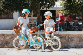 Deux filles de 5 et 6 ans assises à côté de leurs vélos enfant Bebikeen 16 pouces, bleu turquoise et crème avec panier en osier et béquilles. Elles portent un casque assorti.